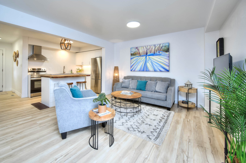 Living room space skillfully staged by Helen's Team, seamlessly integrating a kitchen area and furnished with stylish gray couches.