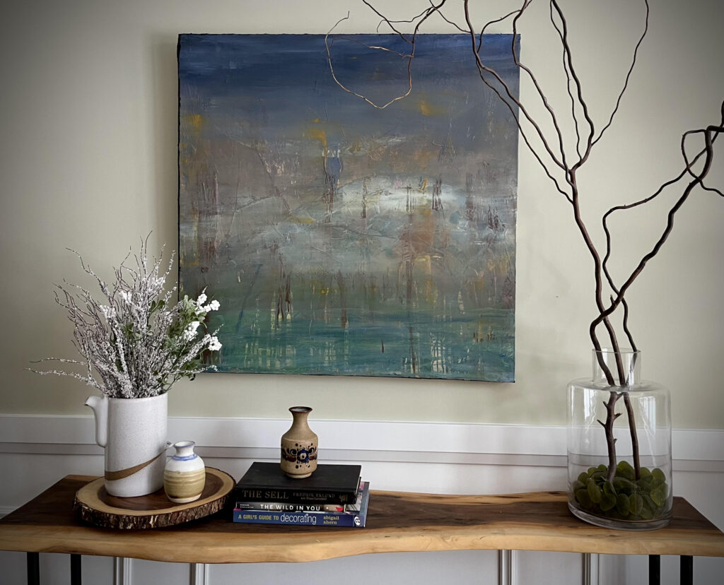 Wooden table with books, vases, and twigs arranged neatly on top, set against a white wall featuring a framed painting.